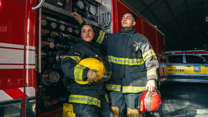 Two firefighters in full gear stand by a firetruck inside a station., tags: bei - pexel